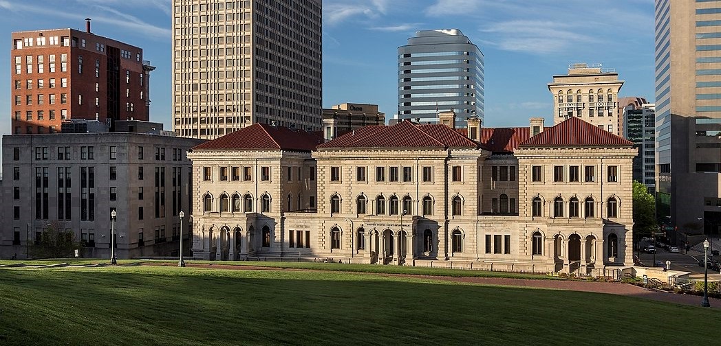 A photo of the Lewis F. Powell, Jr. Courthouse with several downtown Richmond buildings in the background and a grass lawn in the foreground. The building is three stories tall and in the Italianate style with white stone walls and a red tile roof.